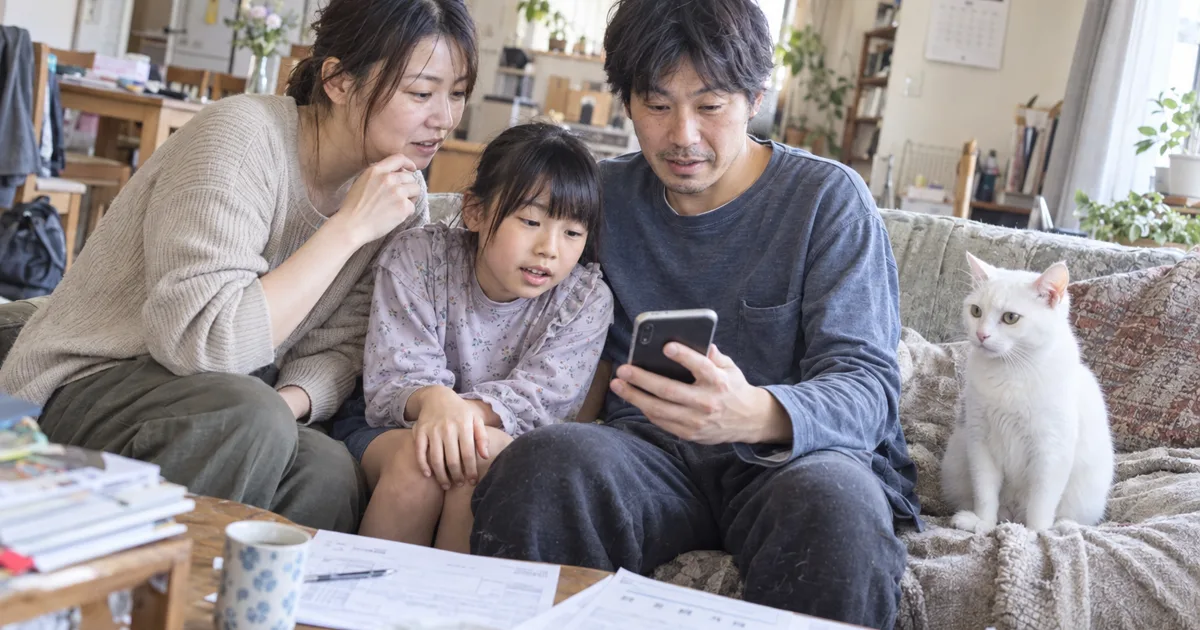 ソファでスマートフォンの料金表を見る3人家族と、隣に座る白猫